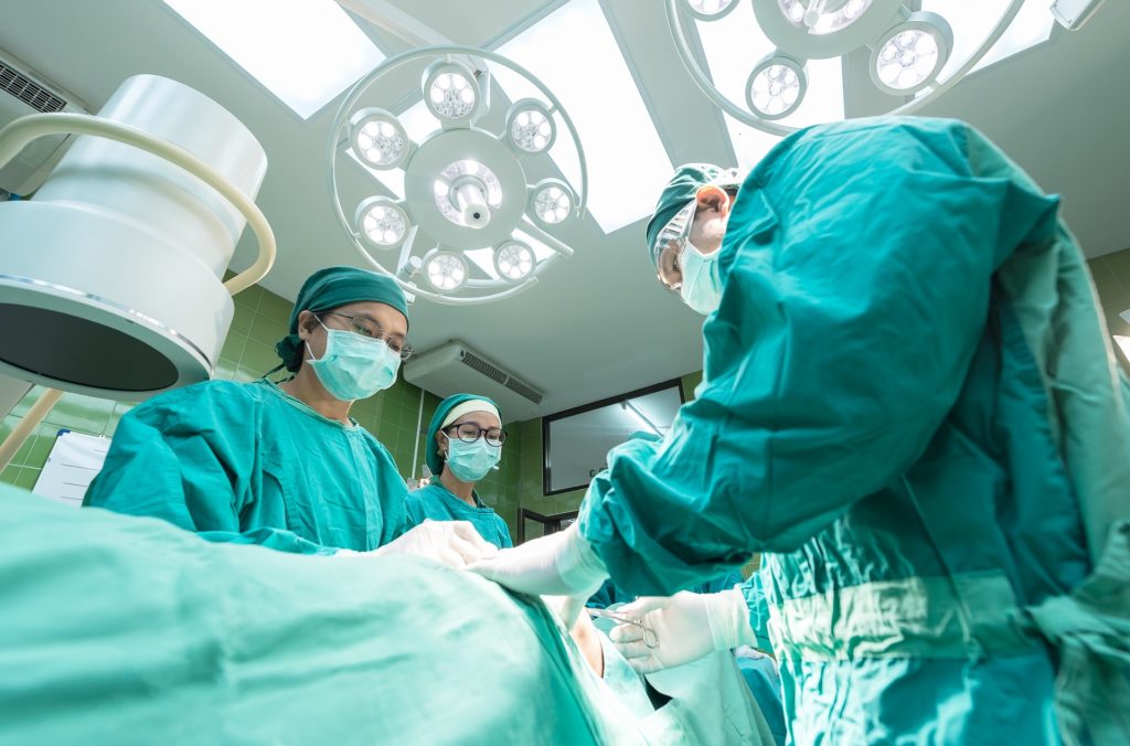 Sala de cirurgia com 3 médicos, vestidos com roupas próprias verdes e máscara, um paciente na mesa de cirurgia.