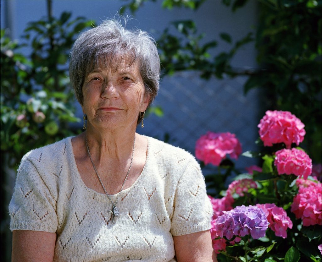 Senhora de cabelos brancos, camiseta bege em frente a algumas flores rosas